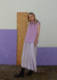 Woman wearing a light purple sleeveless top and pale lilac skirt standing against a textured wall with purple panels.