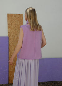 Woman wearing a lilac sleeveless knitted top and skirt standing against a plain wall.